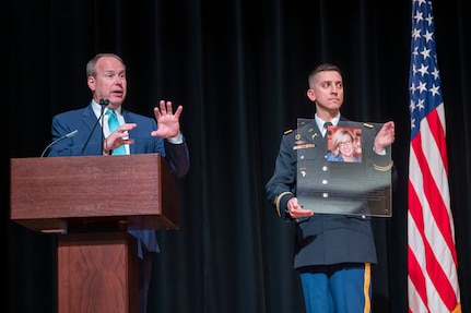 A man in a suit speaks from a microphone lectern next to a man in uniform holding a plaque.