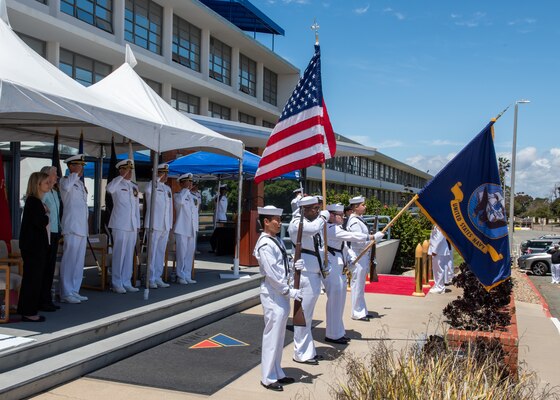 Color Guard holding flags in front of saluting sailors under a white tent.
