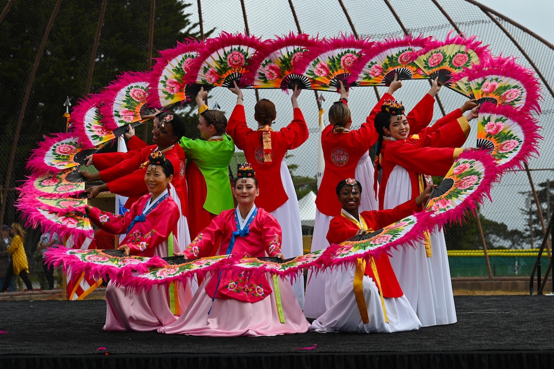 A group of people dressed in Korean traditional clothing  stand in a circle and hold large traditional fans.