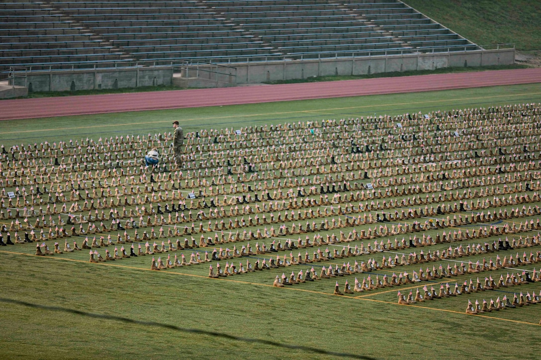 A soldier stands next to a person kneeling in a stadium field filled with military boots.