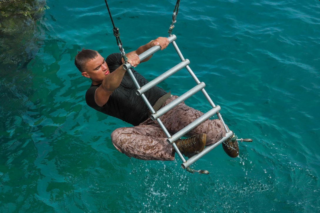 A Marine holds onto a ladder that is hoisted above a body of water.
