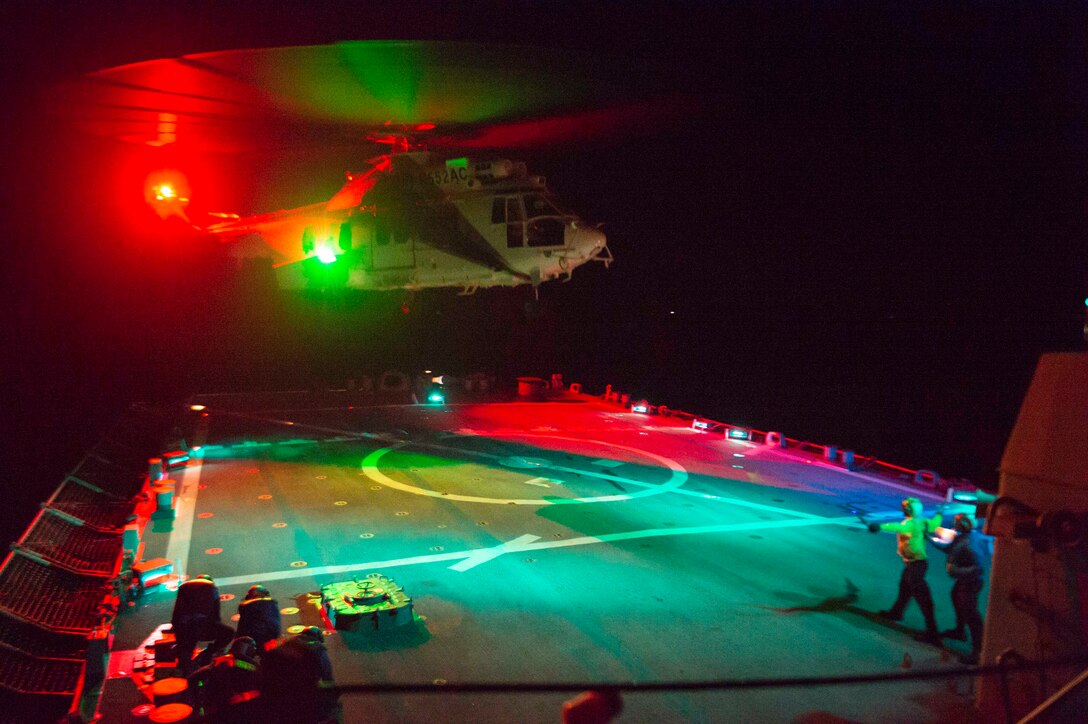 Sailors stand on the flight deck if a ship as a military helicopter lifts off at night.