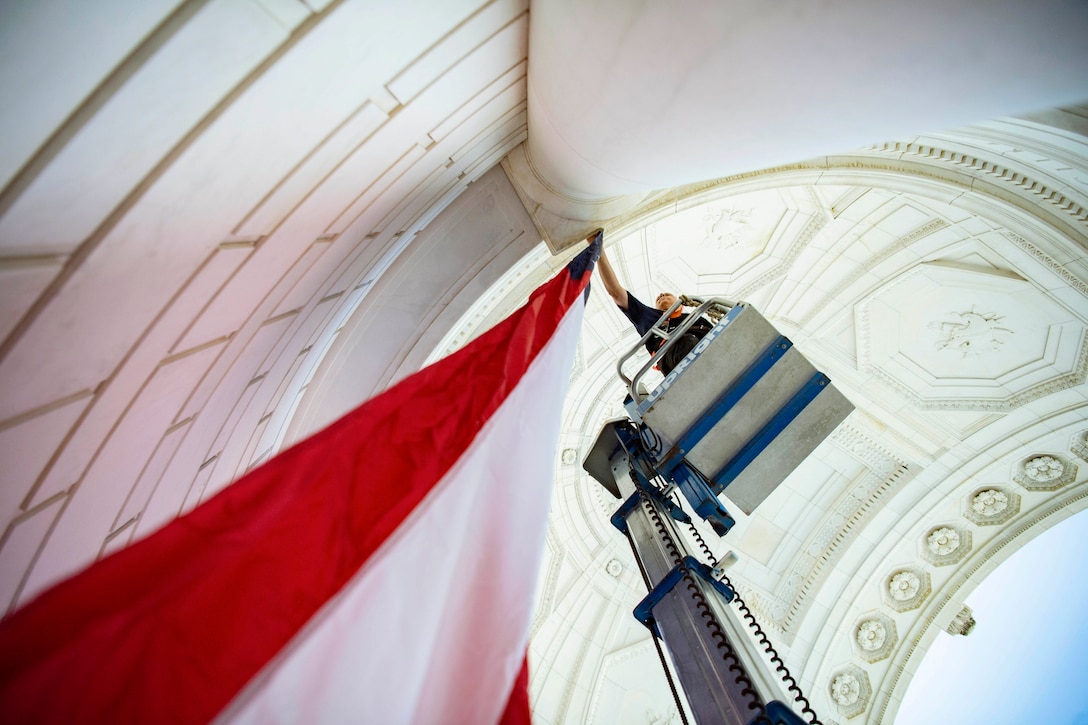 A staff member standing on a lift hangs a flag in a memorial amphitheater.
