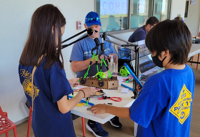 Petty Officer 1st Class Adam Massingil, Pearl Harbor Naval Shipyard and Intermediate Maintenance Facility, center, observes two children, standing at a table, testing a load they built by lifting it with a crane, at the PHNSY demonstration table Onizuka Day of Exploration.