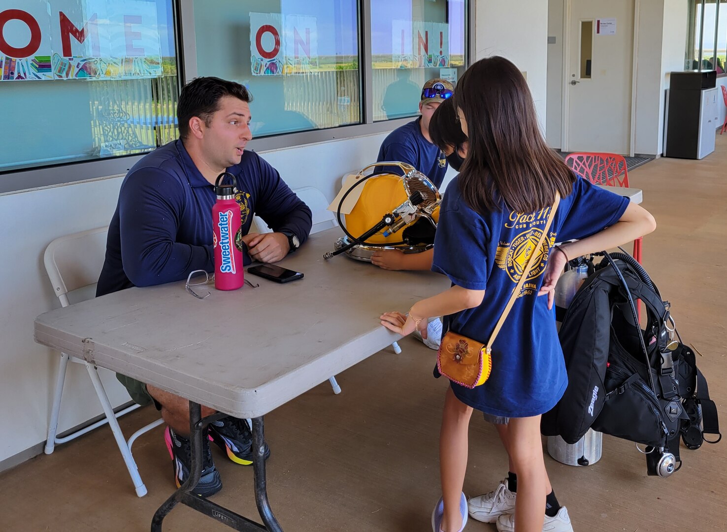 Navy Diver 2nd Class Cameron Duffy, Pearl Harbor Naval Shipyard and Intermediate Maintenance Facility, left, explains how a KM 37 hard hat helmet (diving helmet) works to two children at the shipyard’s demonstration table during the Onizuka Day of Exploration event.