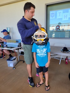 Navy Diver 2nd Class Cameron Duffy, Pearl Harbor Naval Shipyard and Intermediate Maintenance Facility (PHNSY&IMF), left, places a KM 37 hard hat helmet (diving helmet) on a child’s head at the PHNSY & IMF demonstration table during the Onizuka Day of Exploration.