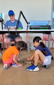 Two child sit on the floor building a load to test on a crane attached to a table as Petty Officer 1st Class Adam Massingil observes from a seat at the table.