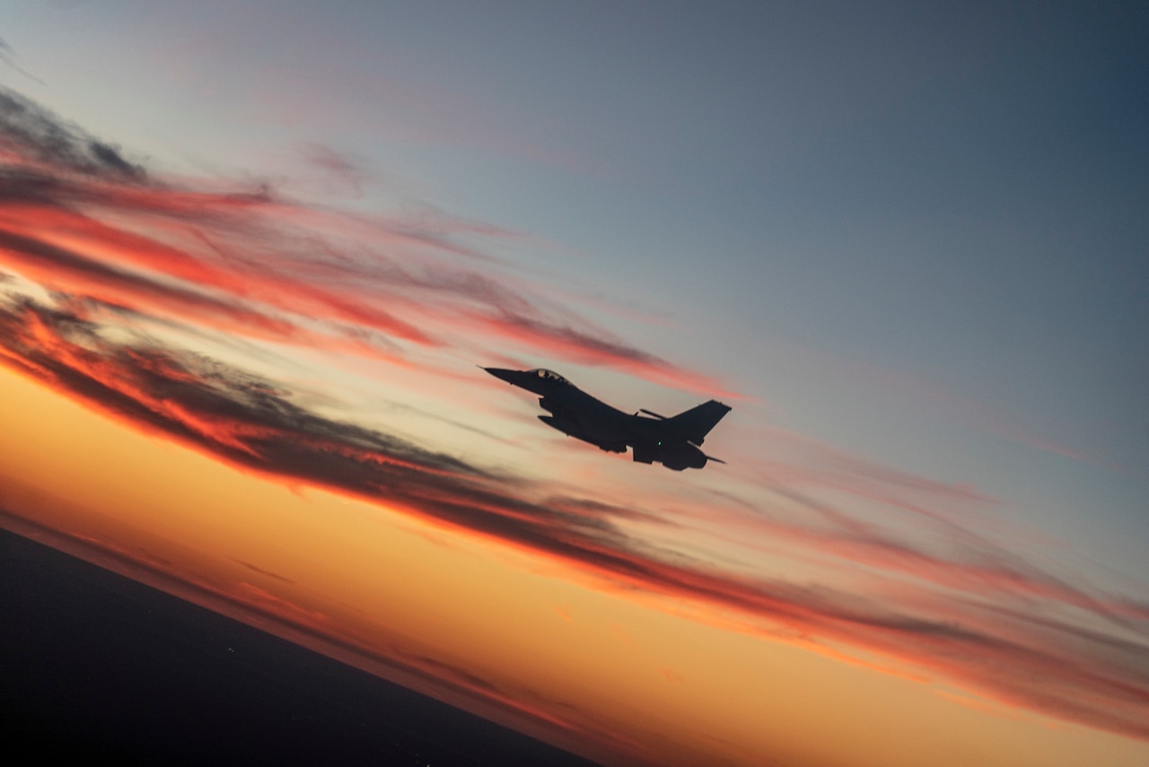 A fighter jet flies in silhouette against a blue and orange sky.
