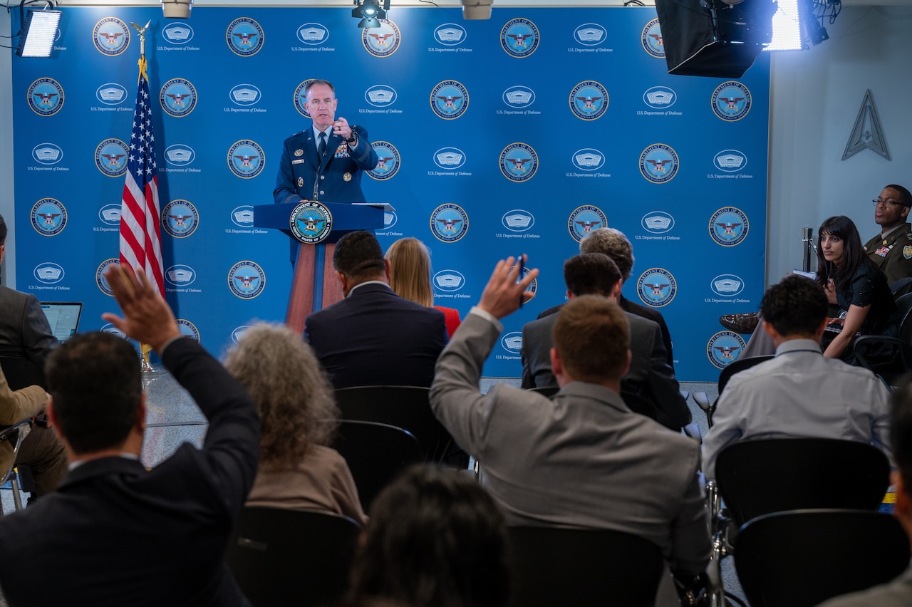 A man stands behind a lectern and points to others who are seated in the room.