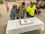 Photo is of three people, one woman, one man in military uniform and another man cutting a big sheet cake.