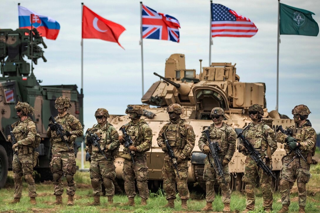 Soldiers stand side by side holding weapons in front of tanks while multinational flags wave behind them.