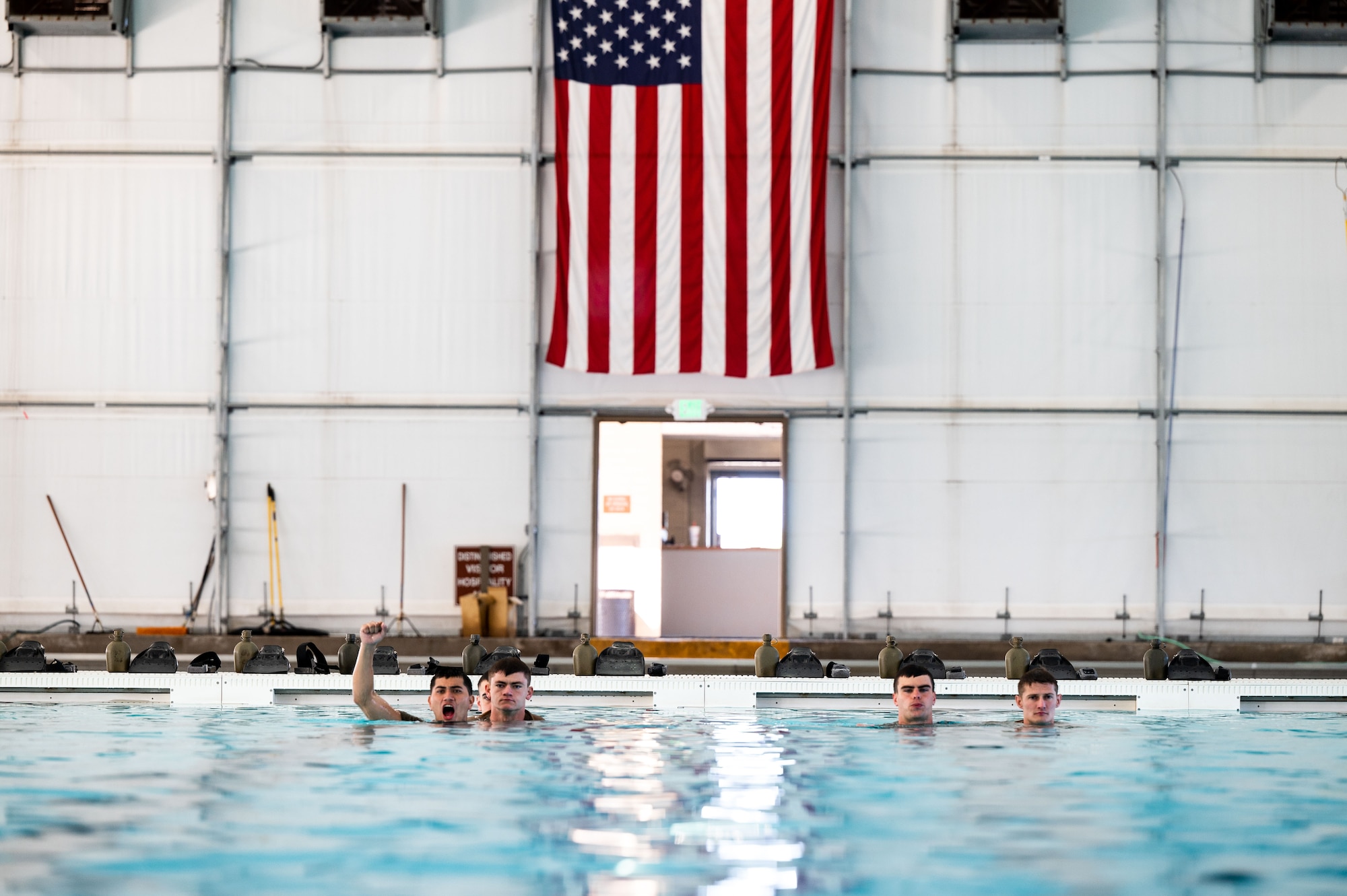 trainees line up in the pool with American flag in the background. One trainee yells and lifts his hand to show he's ready to dive