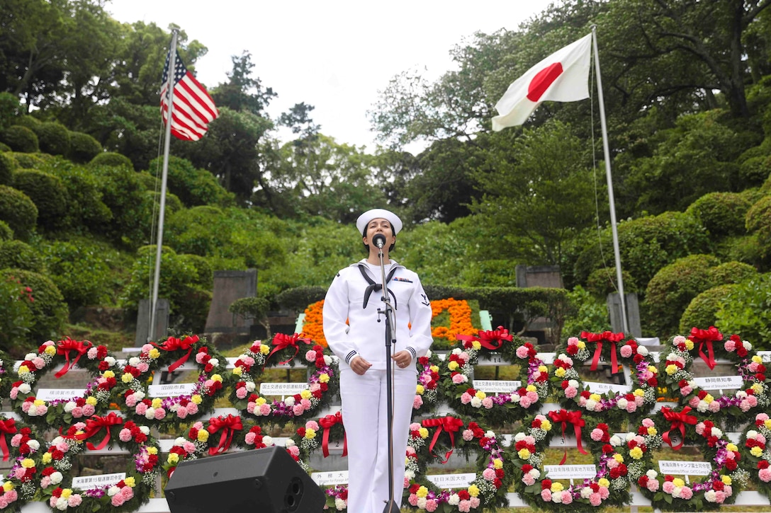 A sailor sings into a microphone in front of rows of wreaths and two flag poles.
