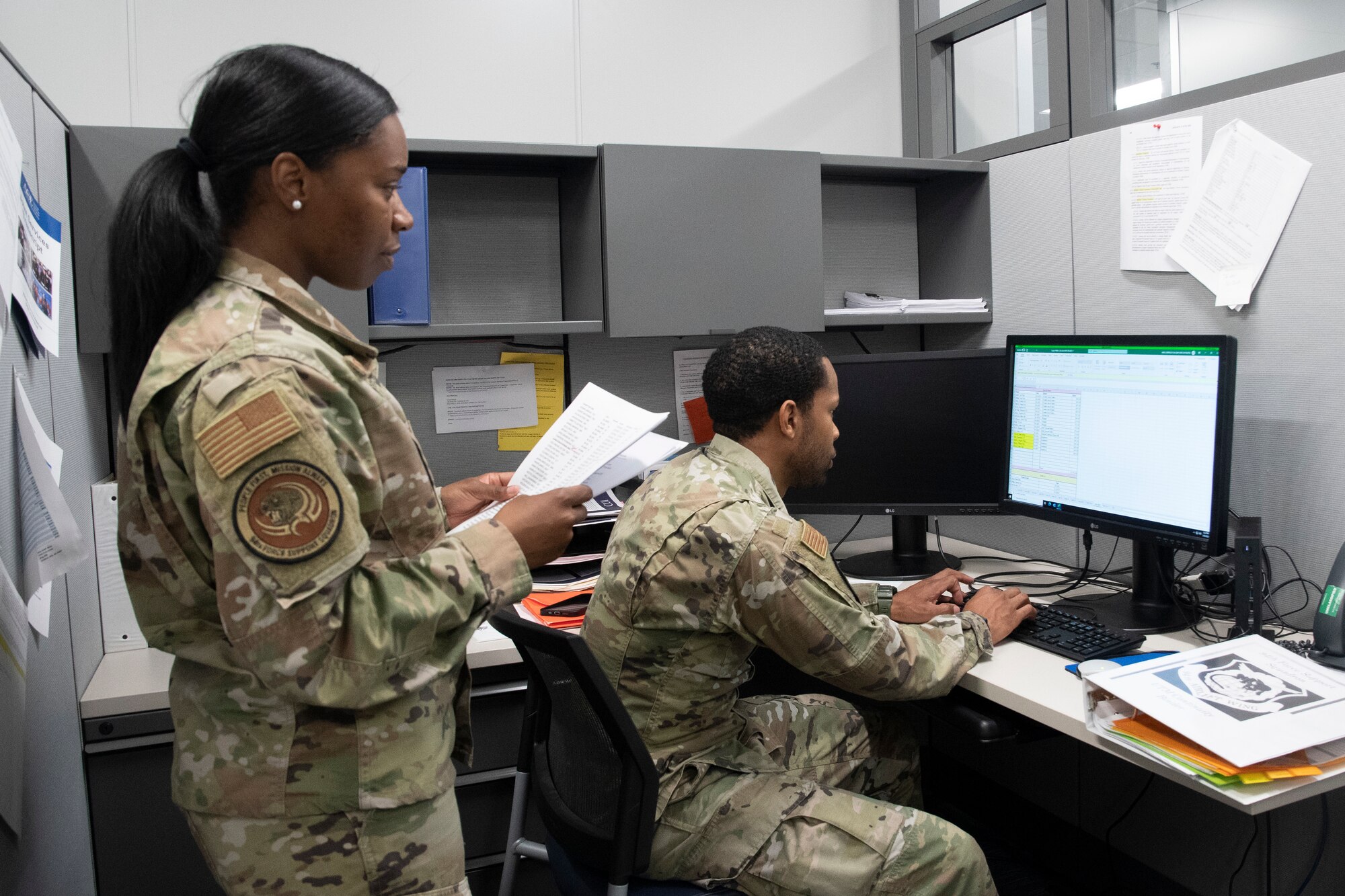 A man and a woman stand and sit, respectively, behind a desk, working on lodging information together.