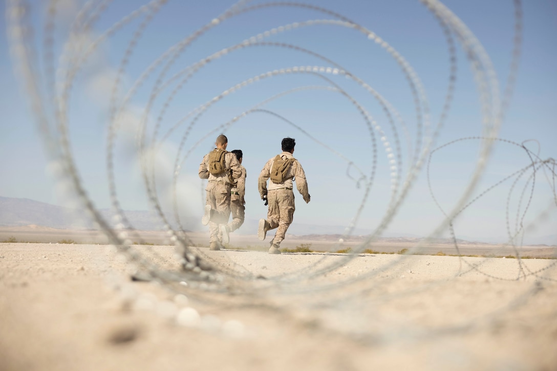 Three Marines run in a desert-like area as seen through barbed wire.