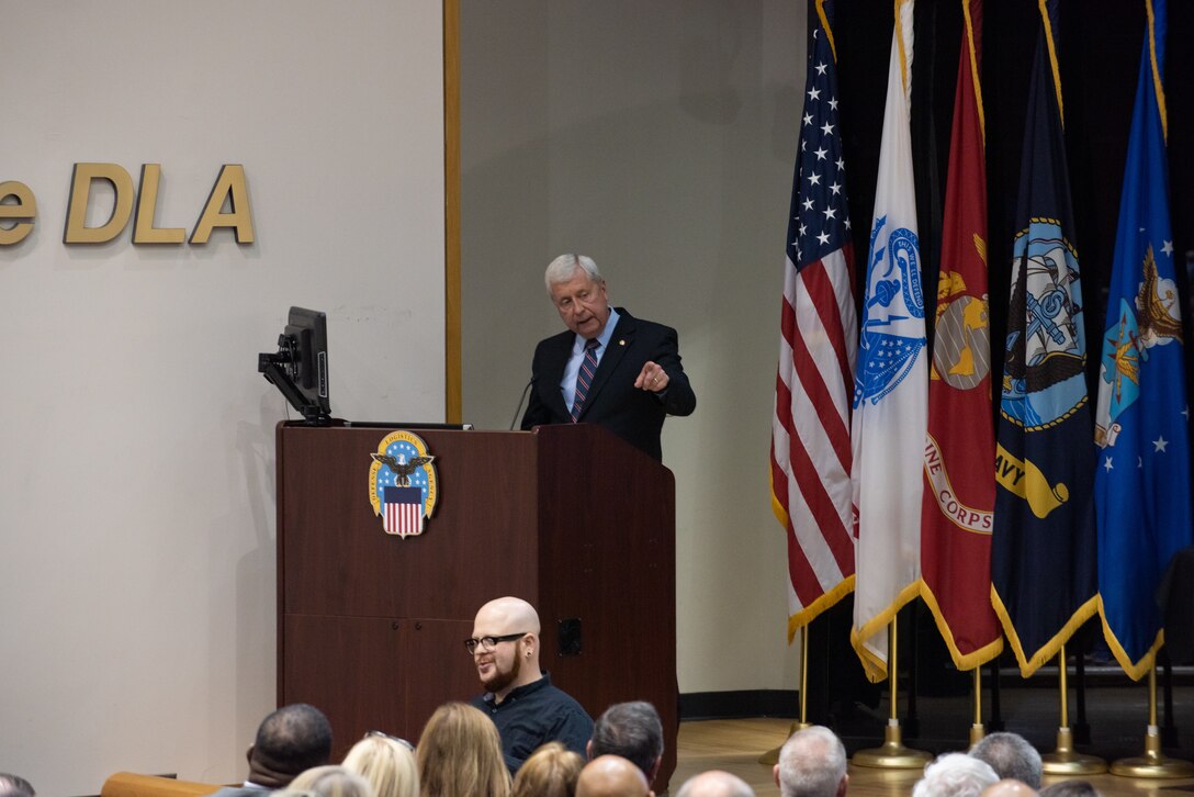 A light skinned white haired man speaks at a podium in an auditorium. He wears a dark suit.