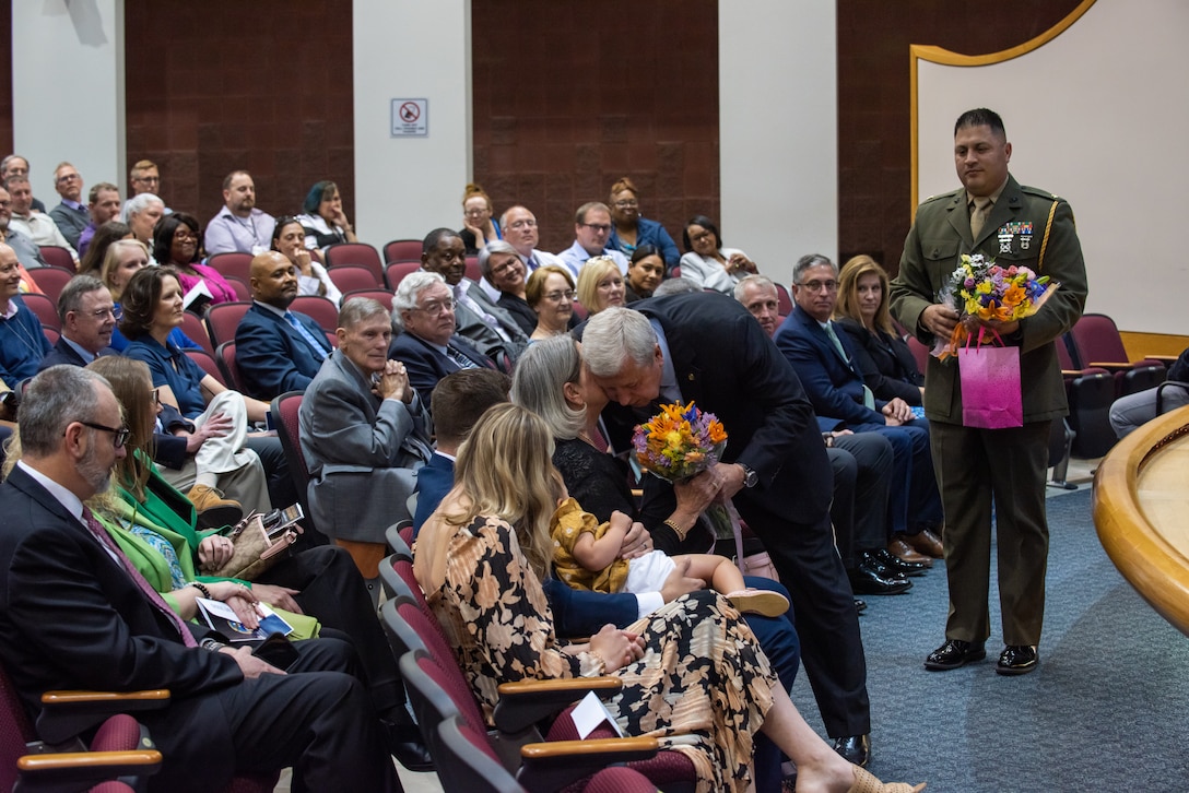 A light skinned white haired man gives flowers to a gray haired woman seated in an auditorium full of people.