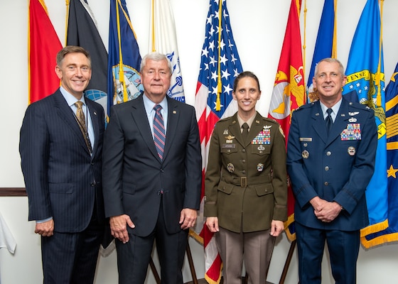 Three men and one woman smile for the camera as they stand in front of a military flag display. The woman wears a Army olive dress uniform, the man on the far right wears a Air Force blue dress uniform and the two men to the left wear dark suits. All are light skinned.