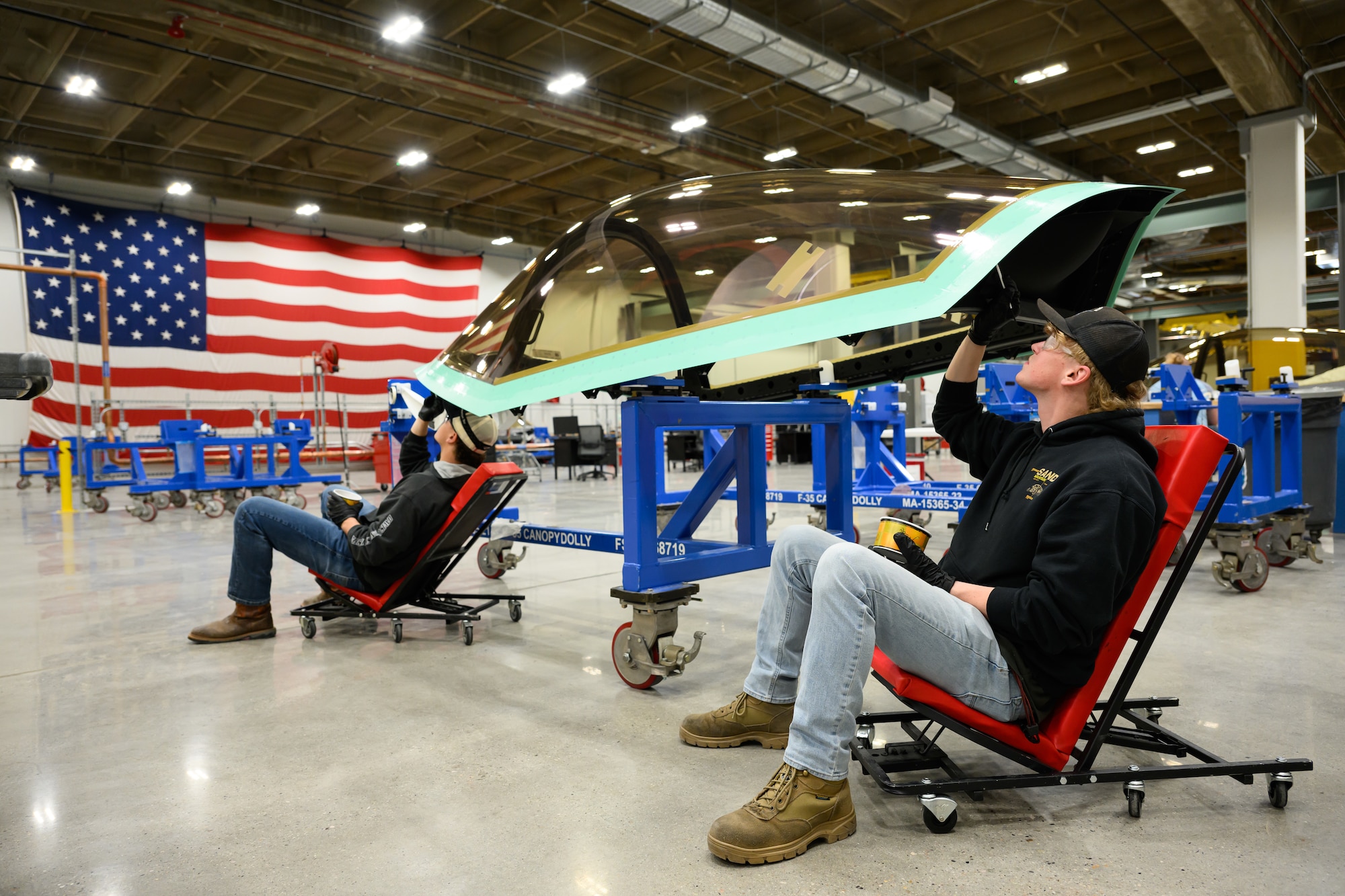 Two men sit on a rolling chair while working on an F-35 canopy