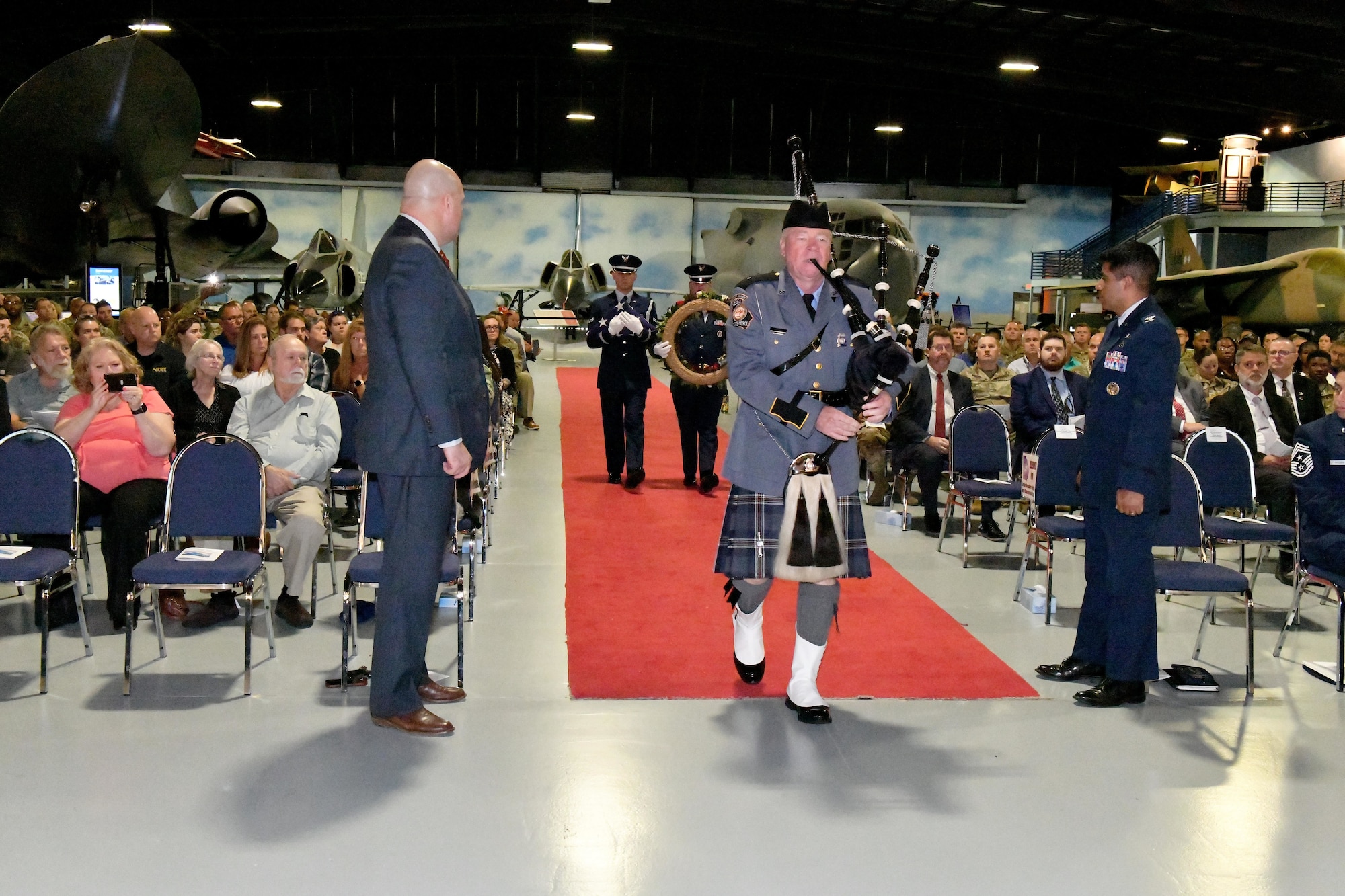 Man walks down aisle playing bagpipes while surrounded by a large group sitting down