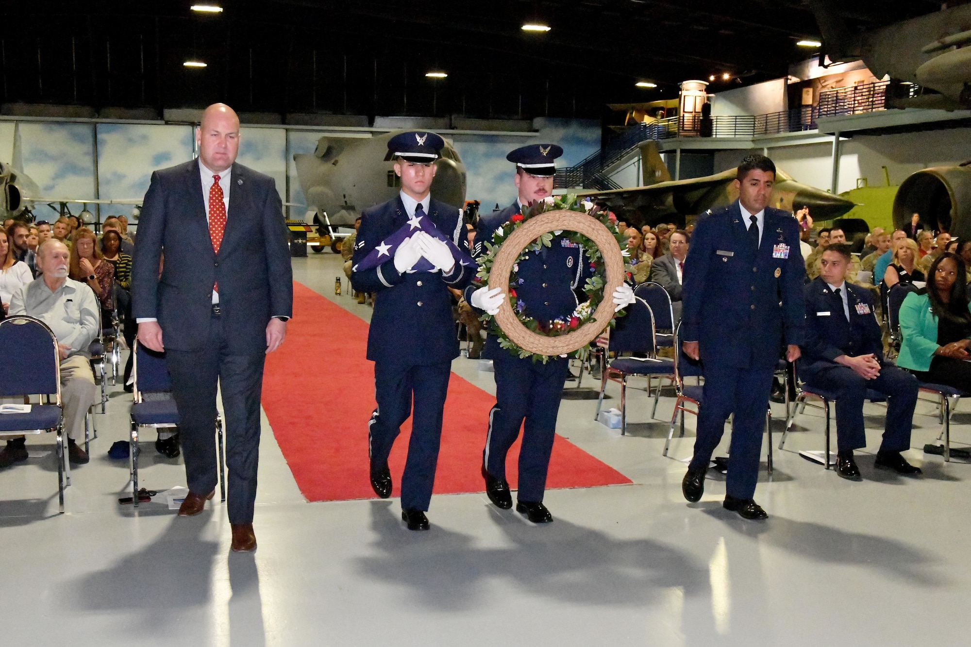A group of three men in military uniforms, with one man in a suit, walking down a runway. One man is holding a wreath.
