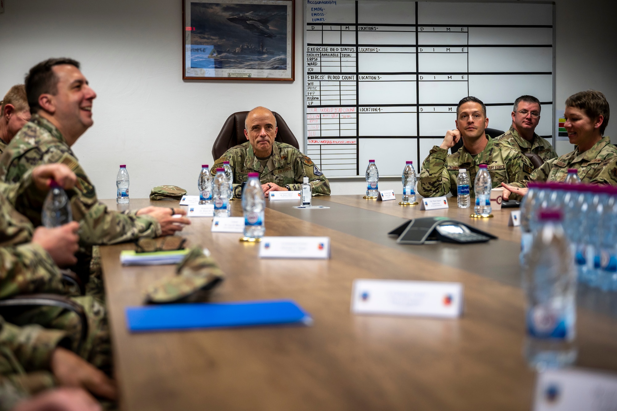 Group of military members sit at a desk listening to a brief.