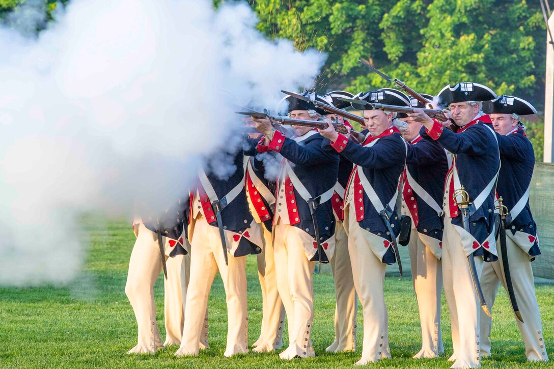 Soldiers stand in Revolutionary War-era attire in formation while firing weapons as smoke surrounds them.