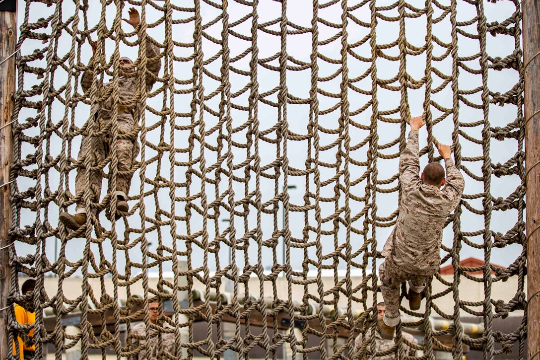 Two recruits climb a rope wall while others watch.