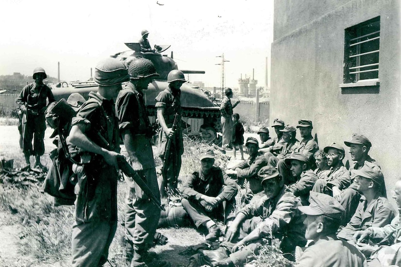 Several men stand over several other men who are seated and looking at them. A tank is in the background.