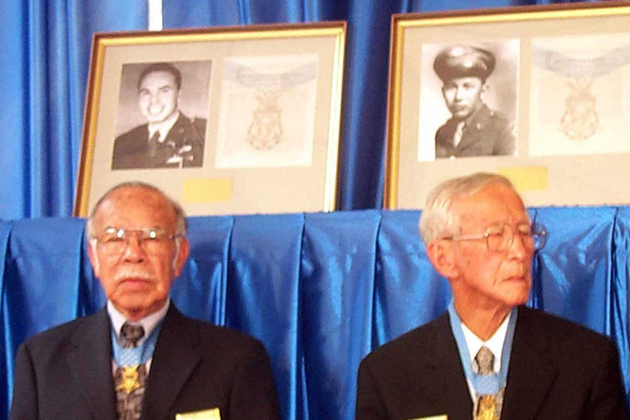 Two men with medals around their necks pose in front of older photos of themselves.