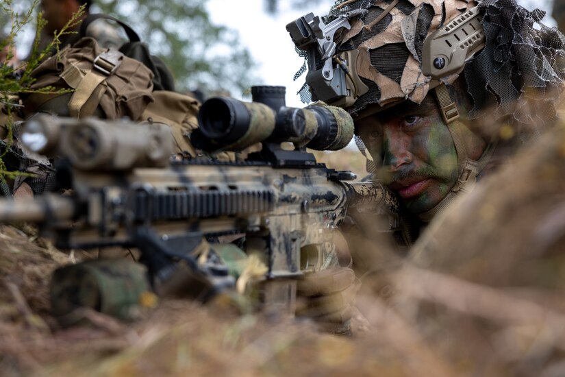 A Marine laying on the ground in camouflage aims a weapon.