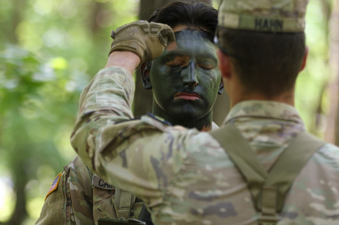 A soldier puts green paint on a fellow soldier’s forehead as they face each other in the woods.