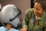 U.S. Air Force Lt. Col. (Ret.) Shawna Rochelle Kimbrell, the first Black female pilot in U.S. Air Force history, helps a child try on a helmet from a fighter jet during the annual Sisters of the Skies, Girls Rock Wings tour, at the 140th Wing, Buckley Space Force Base, Aurora, Colorado, May 20, 2023. SOS is an organization of professional Black female pilots who are committed to supporting future Black aviators through mentorship, professional development, outreach, and scholarship.  (U.S. Air National Guard photo by Tech. Sgt. Chance Johnson)