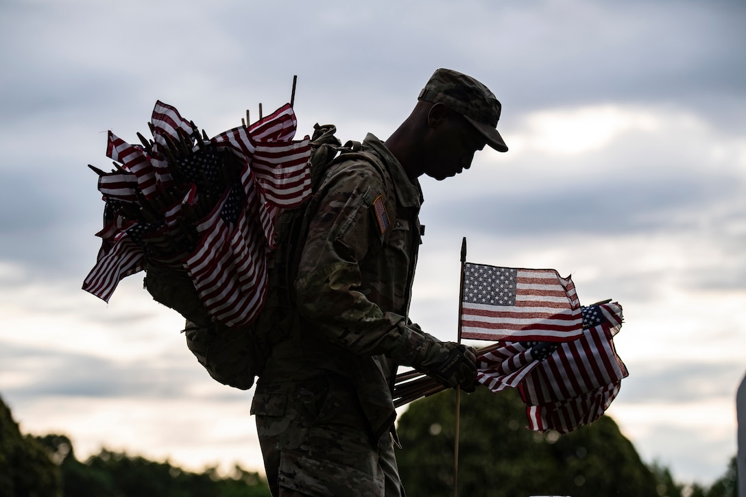 A soldier in silhouette carries American flags in his hands and in a backpack.