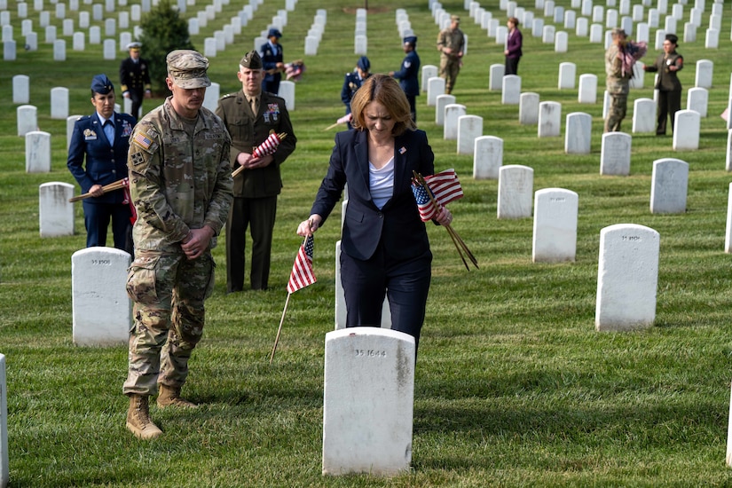Deputy Defense Secretary Kathleen H. Hicks holds American flags and looks at a headstone in a cemetery while staff members look on.