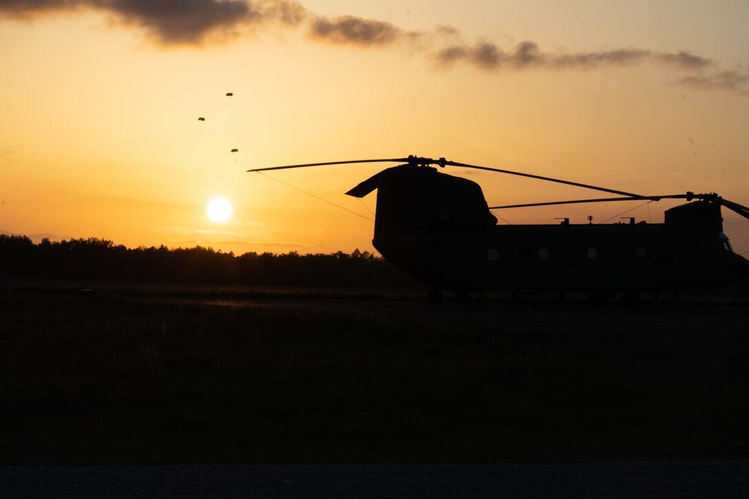 Paratroopers are seen in the distance and an aircraft in the foreground all in silhouette against a yellow sunset.