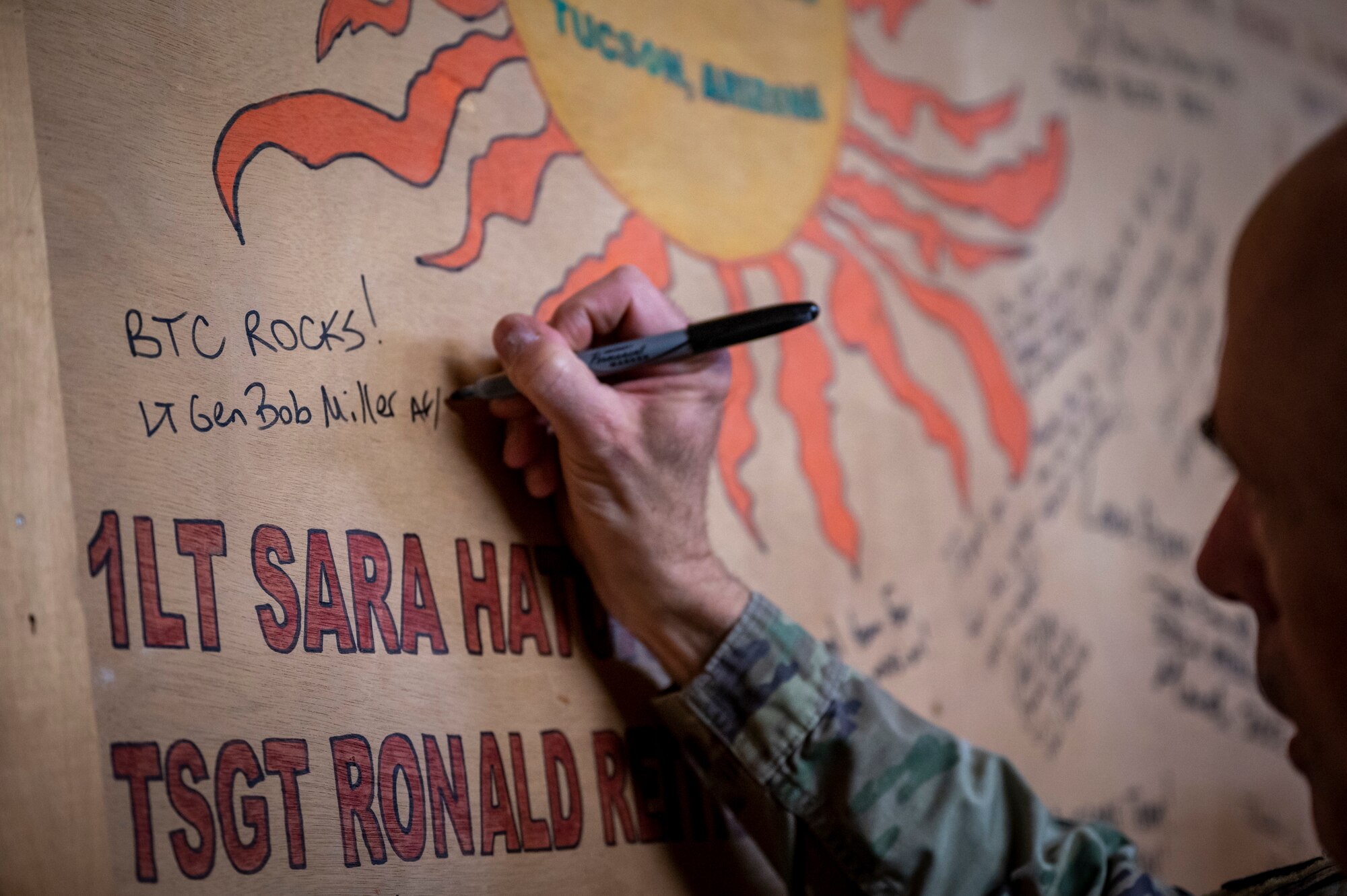 A military member signs a wooden wall covered in autographs.