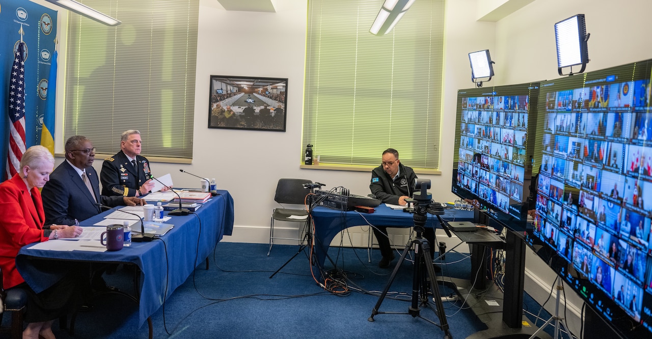 Three people sit behind a long table and look at two large monitors.