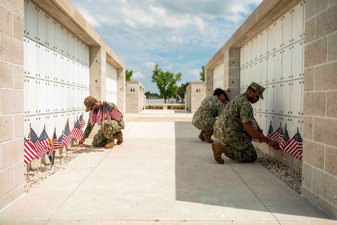 Three sailors place small flags in front of structures at a cemetery.