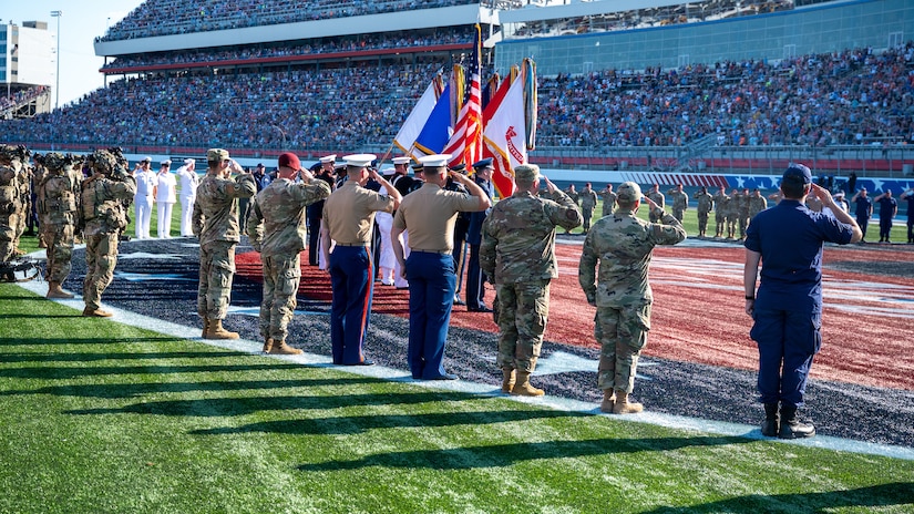 Several people stand on the rim of a large logo on a field. They all salute toward a stadium of people.