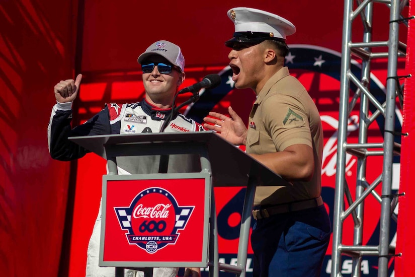 A man in uniform yells into a lectern microphone as a man in racecar gear smiles and gives a thumbs-up.