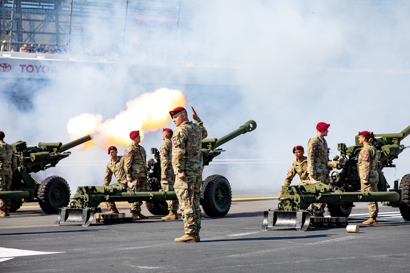 Crews man three howitzers on a track. One of the howitzers just fired, leaving behind a smoke plume.