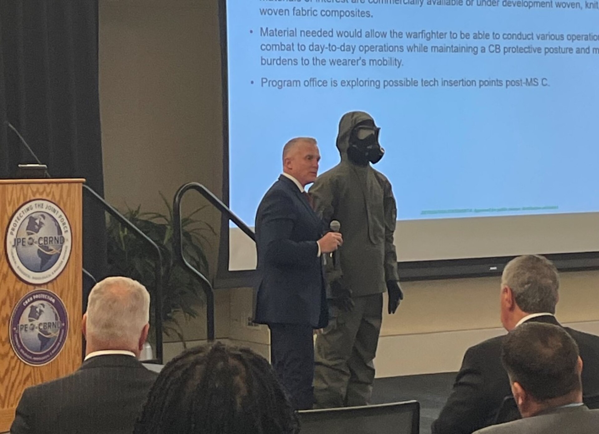 Mr. Darryl Colvin, JPEO-CBRND, speaks to an audience as he stands next to a mannequin wearing the UIPE-FOS CBRN protective gear.