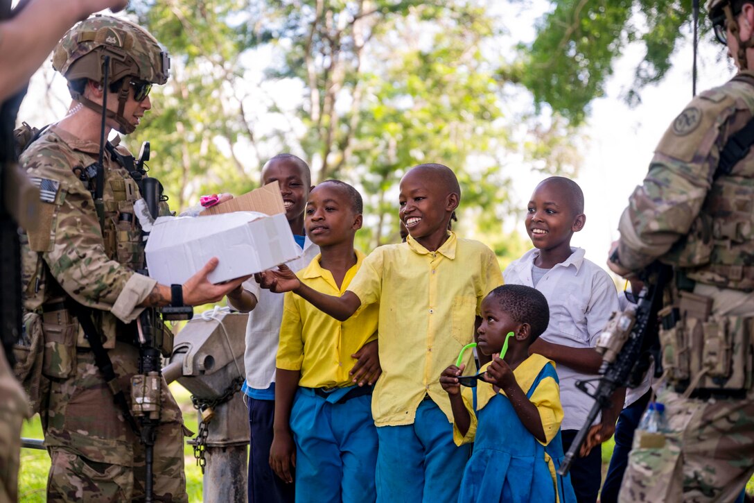 Children smile as a soldier hands out supplies from a box with greenery in the background.