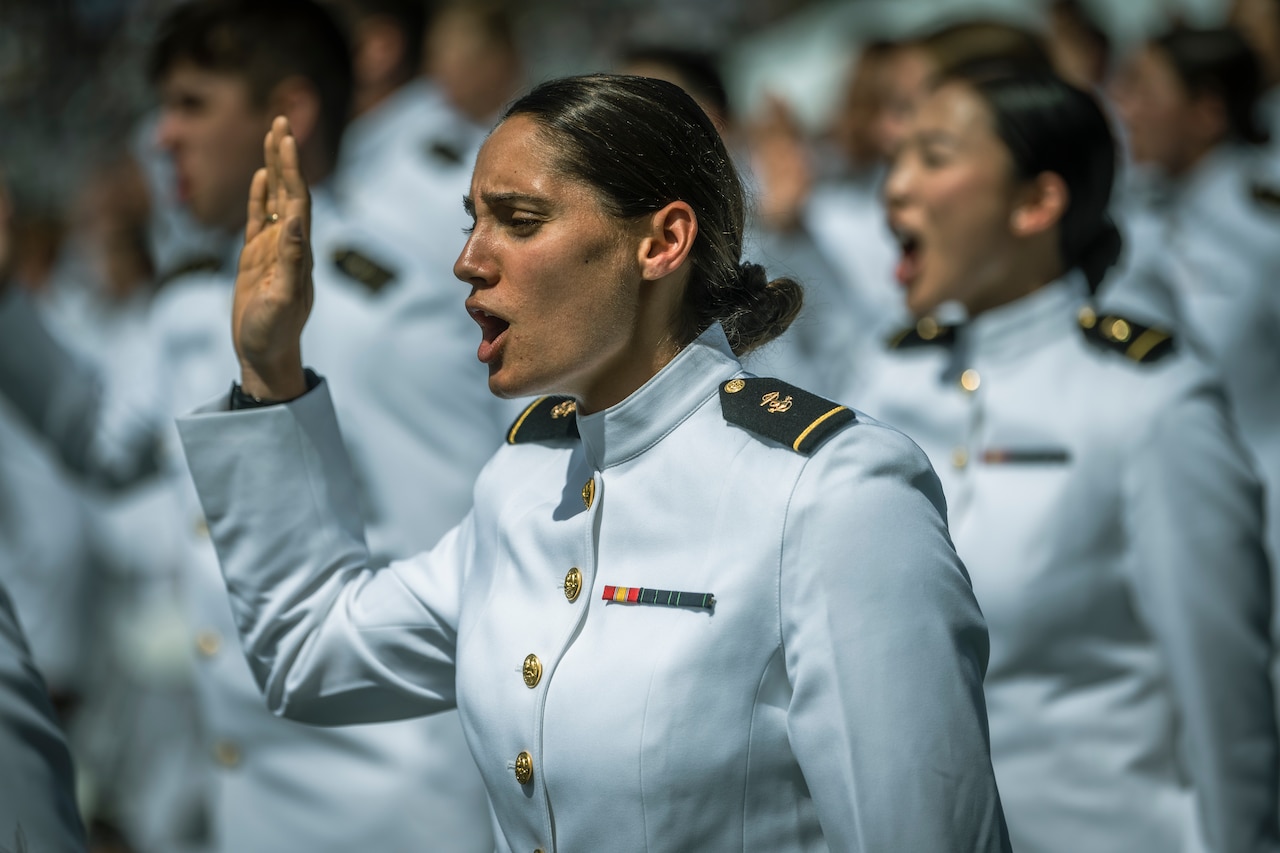 A woman in uniform stands at attention with her right hand raised.