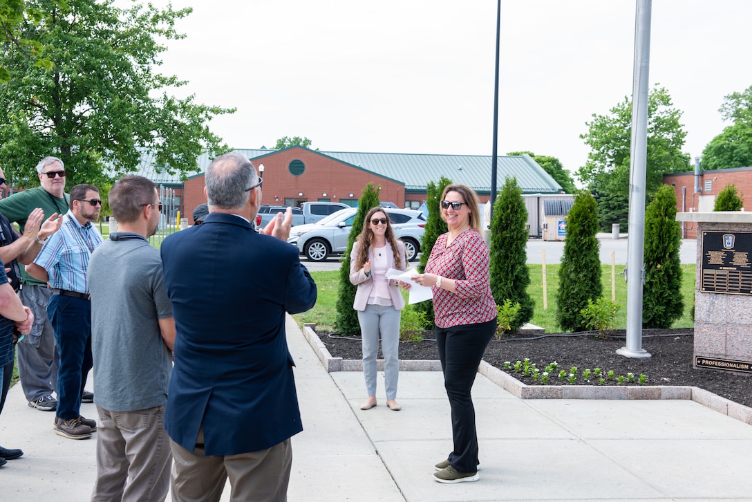 A group of people stand near a memorial at a memorial plaza with flagpoles and green triangle evergreen trees. It is outside and sunny. They are smiling and clapping.