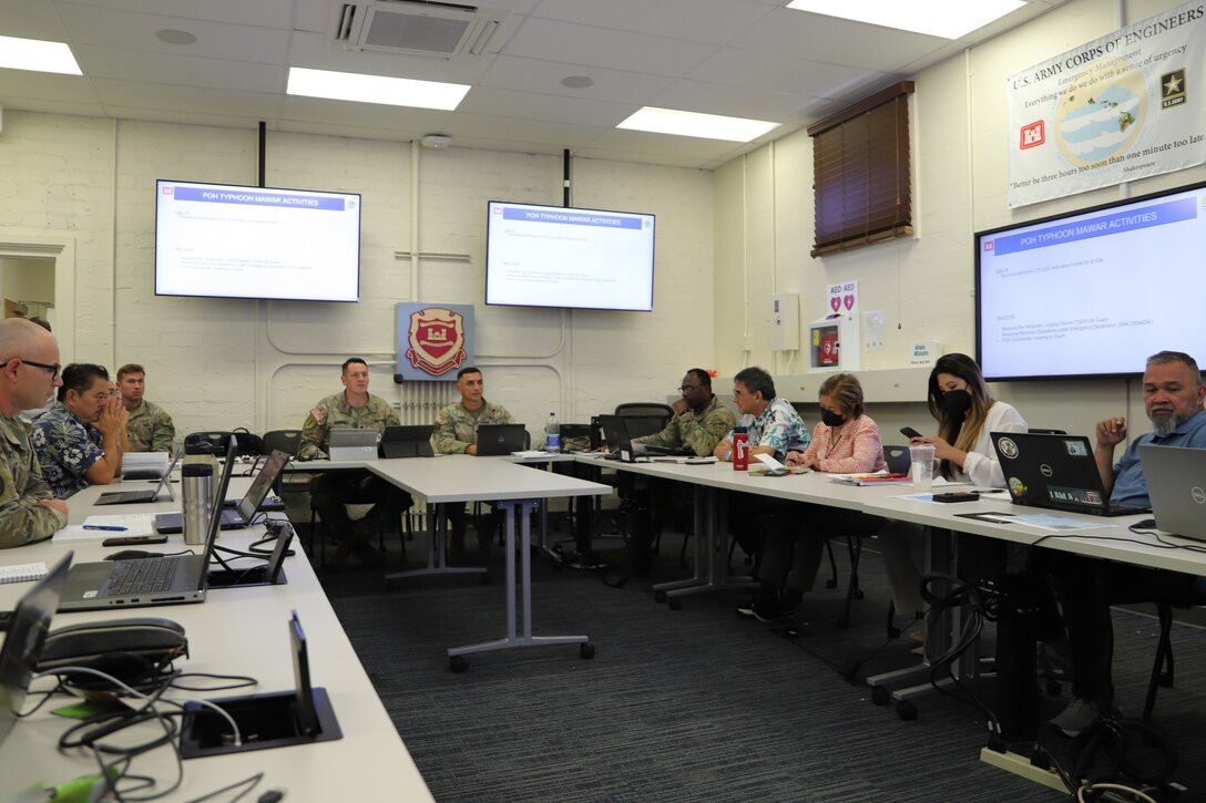 A group of people sit in a white wall command center with monitors on the wall. They are discussing final strategies for teams support Typhoon Mawar disaster relief.