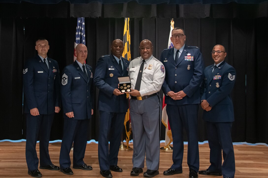 From left, U.S. Air Force Staff Sgt. Zachary Bozell, 316th Security Forces Squadron military working dog trainer, Chief Master Sgt. Jackson A. Helzer, 316th Wing command chief, Col. Todd E. Randolph, 316th Wing and installation commander at Joint Base Andrews, Md., Malik Aziz, Prince George's County chief of police, Col. Michael Morales, 316th Security Forces Group commander, and Tech. Sgt. Amaya, Tech. Sgt. Manuel Amaya, 316th Security Support Squadron combat arms noncommissioned officer in charge, participate in a ceremony at Prince George's County Police Department Headquarters in Upper Marlboro, Md., May 25, 2023. PGPD presented a gift of appreciation as a thank you to military members stationed at JB Andrews and in celebration of Military Appreciation Month. (U.S. Air Force photo by Senior Airman Daekwon Stith)