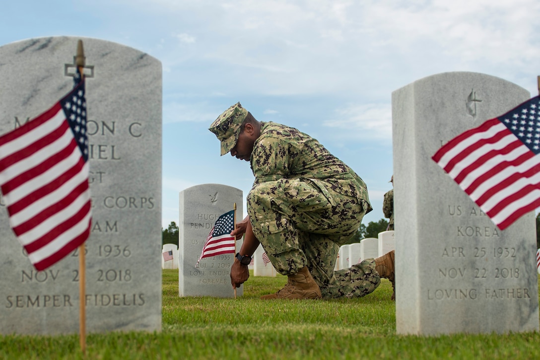 A sailor kneels to place a flag at a headstone, seen between two other headstones with flags.