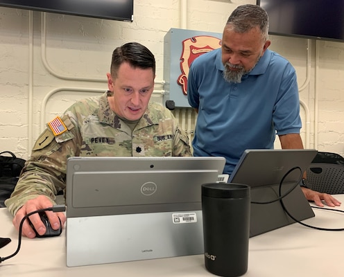 Two men, one wearing an Army uniform and the other wearing a blue polo shirt, sit in front of two silver laptops to review final deployment strategies.